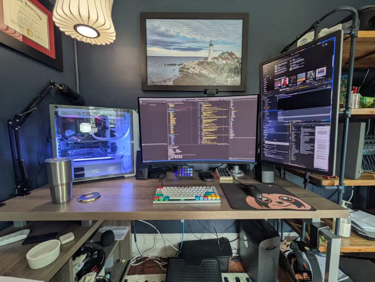 A home-office desk with three monitors showing code and terminals, a custom water-cooled PC with a glass side panel glowing blue, and a mechanical keyboard on a wooden standing desk. A framed lighthouse photograph and a diploma hang on the slate-blue wall, with pipe-style shelving to the right.