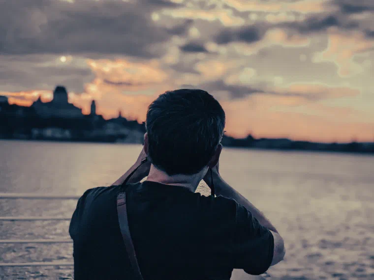Jared Knowles seen from behind, raising a camera to photograph a city skyline across the water at sunset.