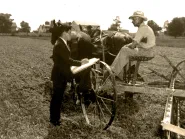 A Census Bureau enumerator in a dark suit stands in a hay field writing in a large ledger as he interviews a farmer in a straw hat seated on a horse-drawn hay rake.